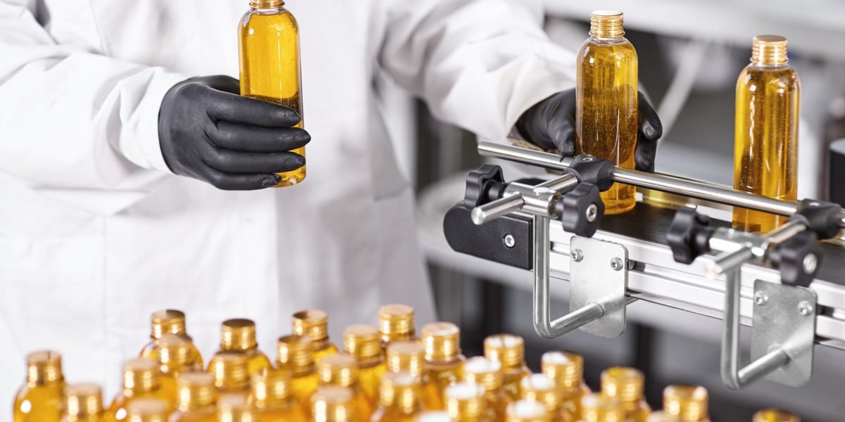Photo of production new kind of cosmetics by professional researcher. Young scientist standing isolated over factory background dressed in gown and black gloves holding bottles with yellow liquid