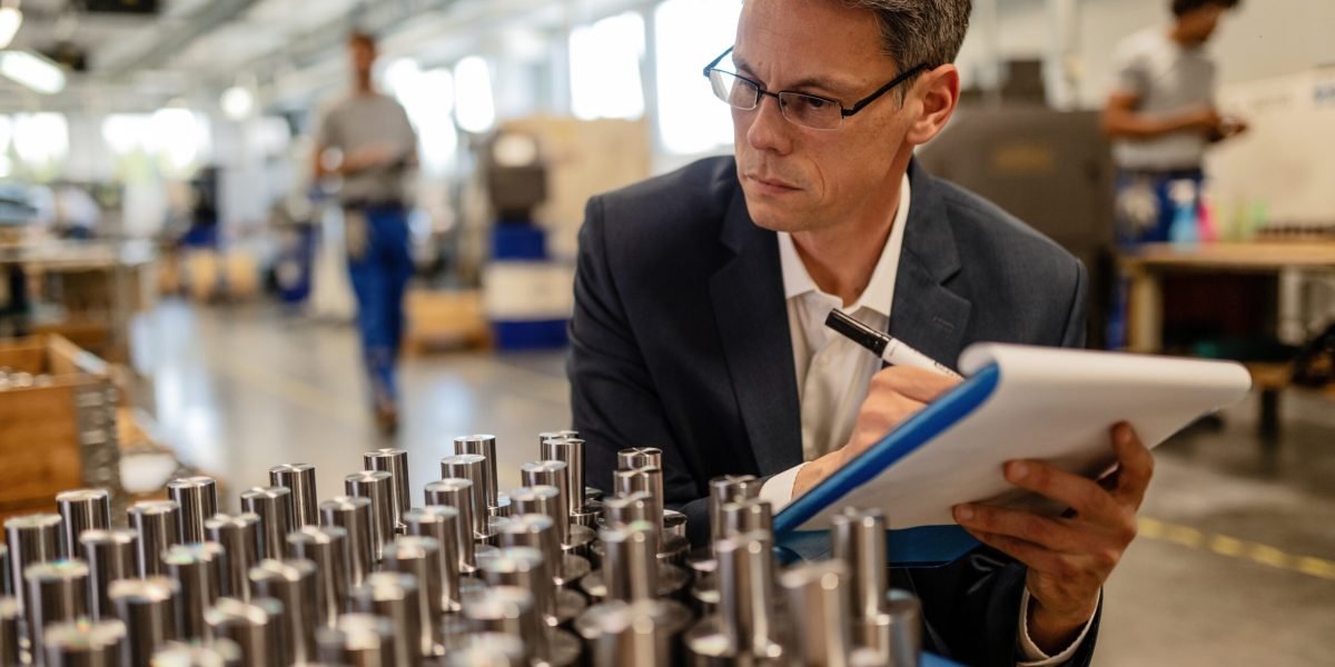 Quality control inspector writing reports while analzying manufactured steel rod cylinders in industrial building.