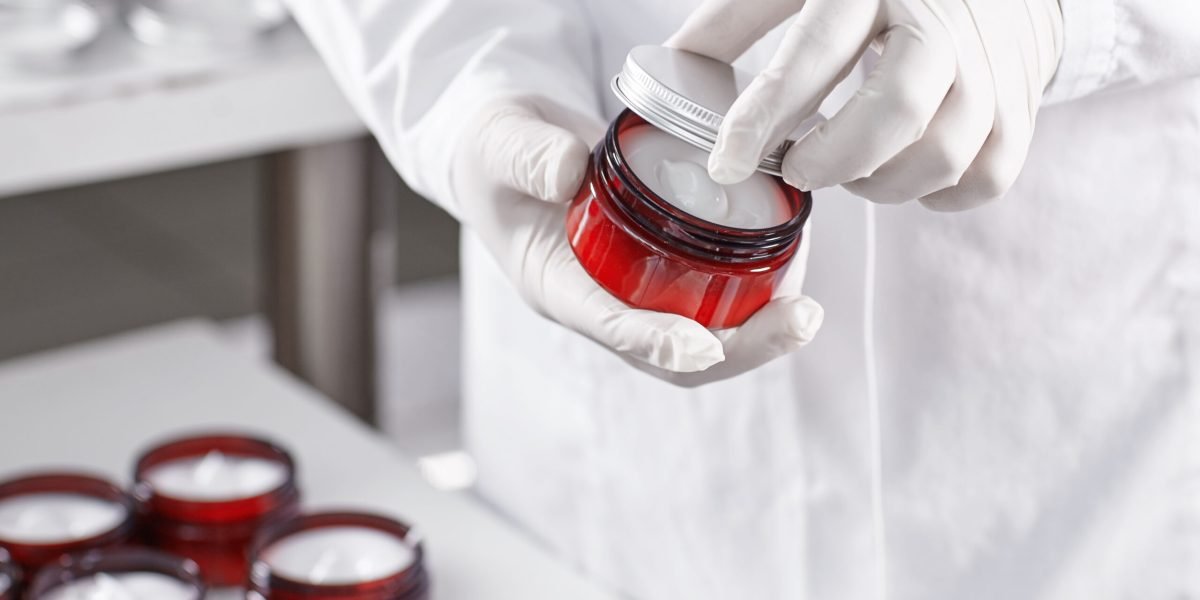 Factory worker in white gown and gloves holding red container with skin care product touching it with finger testing quality. Representing new product on factory. Showing high quality beauty mask